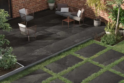 A contemporary patio designed with Brisks' Fantasy Porcelain Paving, featuring two chairs and a small table. The area is enclosed by brick walls and vibrant greenery, including potted plants and a small tree, crafting a cozy outdoor ambiance.