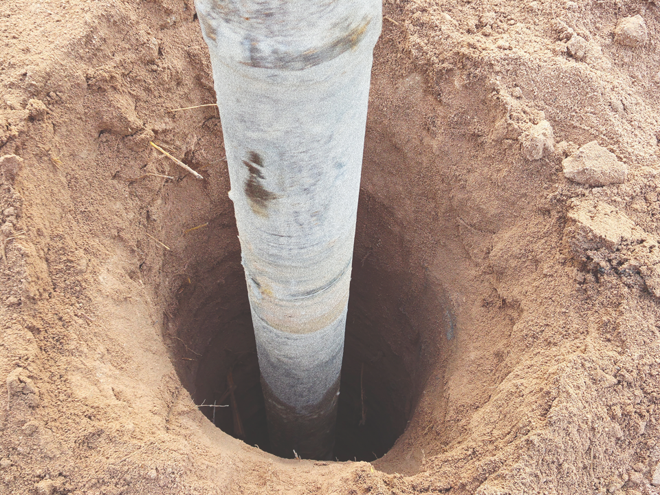 A deep circular hole in the ground with a large gray metallic pole partially buried in the center. The surrounding area is filled with loose, light brown soil, suggesting recent use of Brisks Fence Post Concrete Mix. The image captures a top-down perspective of the scene.