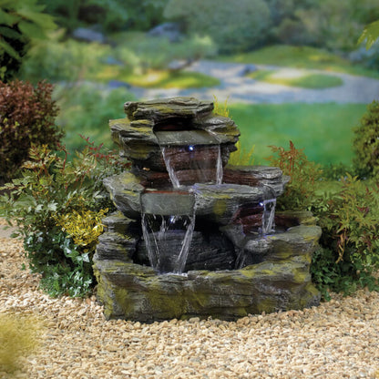 The La Hacienda Garda Falls Water Feature, complete with cascading water and LED lights, is elegantly positioned amidst green plants and shrubs. Behind it lies a grassy garden area with softly blurred greenery, contributing to the serene outdoor ambiance. Pebbles are artfully scattered around the base of this sophisticated garden water feature from La Hacienda.