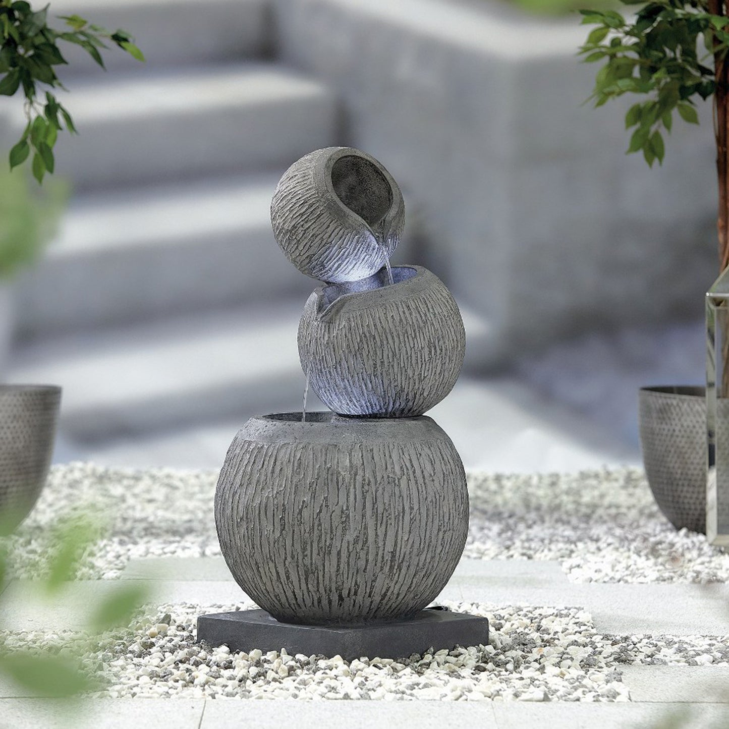 The La Hacienda Illusion Falls (Inc LEDS) Water Feature by La Hacienda features three stacked spherical bowls with water gracefully cascading from top to bottom. Set on a pebble base and surrounded by potted plants, this outdoor water feature is illuminated by LED lights, with the blurred outline of stairs visible in the background.