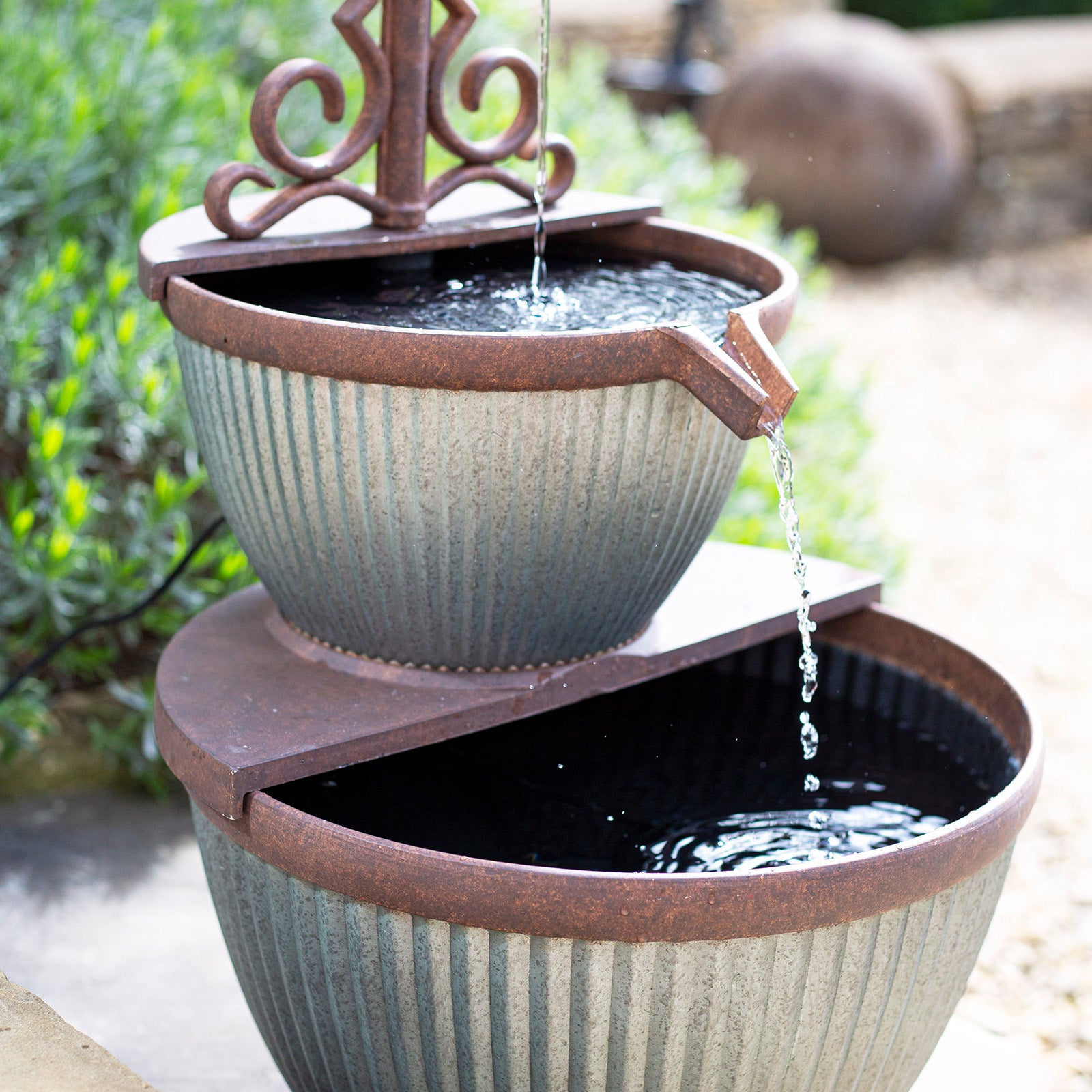 The La Hacienda Irondale Pours Water Feature by La Hacienda is a stunning two-tiered metal garden fountain with a rustic design. It features water cascading from a spout in the upper bowl to the lower bowl, set against a backdrop of greenery and pebbled ground, adding charm to any garden setting.