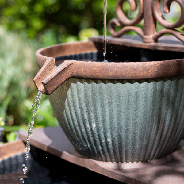 Close-up of a garden water feature with a rustic, corrugated metal basin. Water spills gently from a spout at the edge of the La Hacienda Irondale Pours Water Feature, creating a stream that splashes into a pool below. The background is blurred, showing green foliage, contributing to a serene outdoor setting.