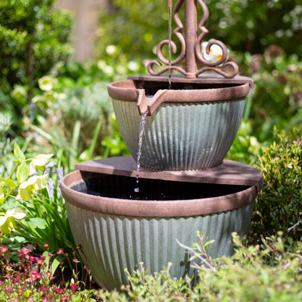 A tiered garden fountain with two blue-green, ribbed ceramic basins is set among lush greenery and blooming flowers. This La Hacienda water feature, the Irondale Pours, elegantly trickles from the upper basin to the lower one, creating a serene and tranquil atmosphere.