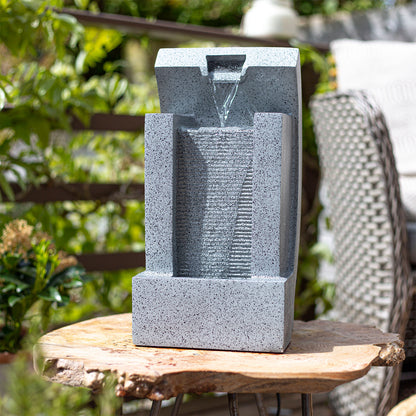 A modern, rectangular tabletop water fountain sits on a wooden surface. The La Hacienda Kiso Column Water Feature, crafted from a gray stone-like material, features water trickling down a textured vertical surface illuminated by LED lights. The background includes greenery and outdoor furniture, suggesting a garden environment.