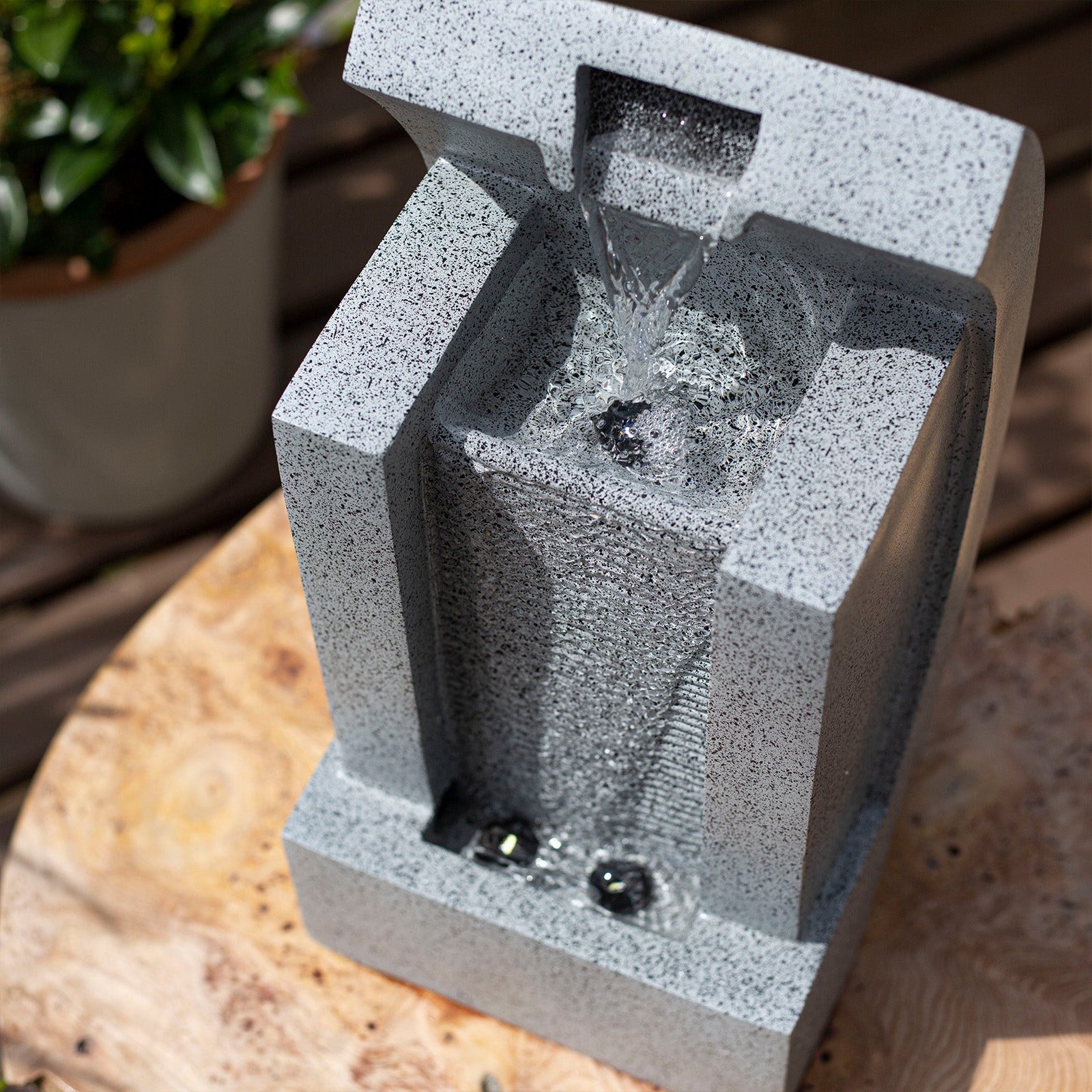 Image of a small, modern tabletop water fountain made of gray, speckled stone. Water flows from the top into a square basin below. Set on a wooden surface in a serene garden environment, this piece, reminiscent of the La Hacienda Kiso Column Water Feature by La Hacienda, is accompanied by soft LED lights and a potted plant in the background.