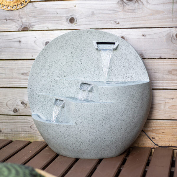 A round, modern tabletop La Hacienda Kyoto Falls Water Feature with three staggered spouts cascading water into shallow basins below, set against a wooden wall backdrop. The fountain features a smooth, light gray modern stone effect surface and is placed on a wooden surface.