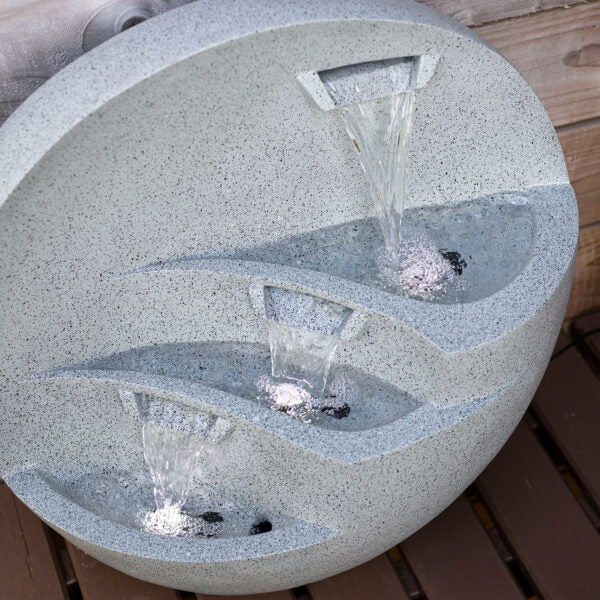 A modern stone-effect, round water fountain with three cascading tiers, the La Hacienda Kyoto Falls Water Feature by La Hacienda adds a touch of garden serenity. Water flows gracefully from the top tier to the middle and bottom tiers, creating a tranquil, aesthetic display on a wooden surface.