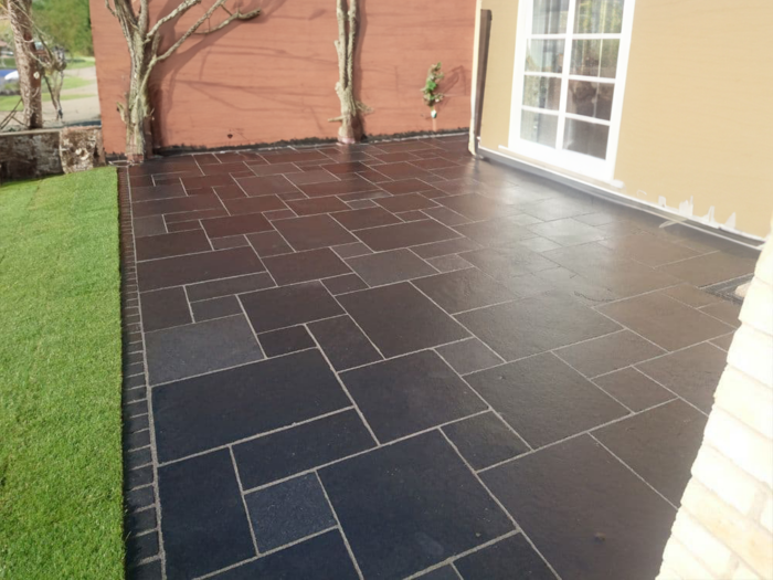 Adjacent to a house with light brown and reddish-brown walls is a patio featuring the Kadappa Black Limestone Patio Pack by Brisks. The dark, rectangular paving slabs are versatile and bordered by a small patch of green grass, appearing freshly cleaned.