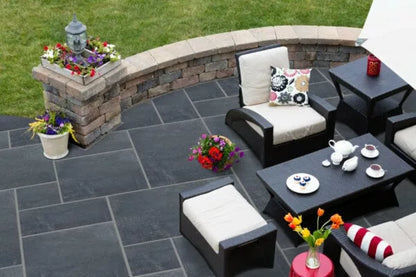 A patio featuring the versatile Kadappa Black Limestone Patio Pack from Brisks beautifully displays a wicker furniture set with chairs and a table. Teacups, a teapot, and pastries are placed on top. Potted flowers and a stone wall adorned with floral arrangements frame this charming space, known for the low water absorption qualities of Brisks' limestone.