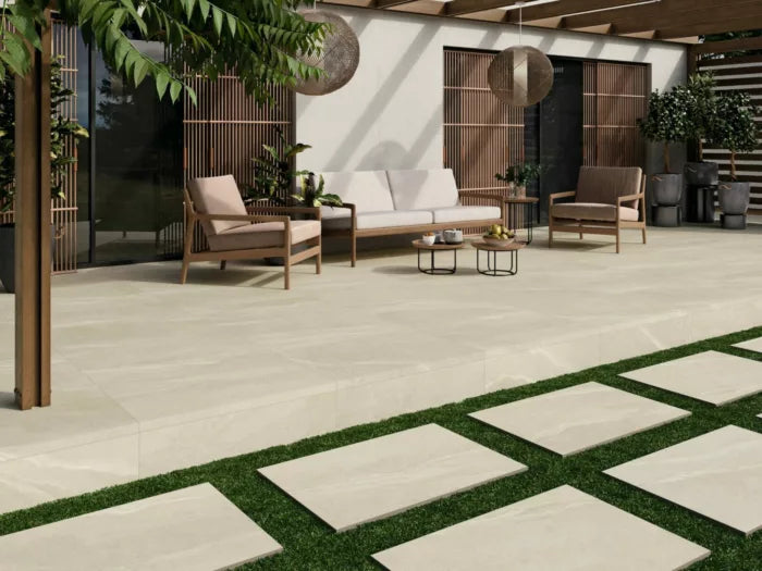 A modern outdoor patio with Brisks Kiara Ivory Porcelain Paving, featuring a white sofa, two armchairs, and small round tables. Wooden paneling and plants decorate the wall, while stone slabs lead across the grass. A pergola with hanging lights adds shade to this chic setting.