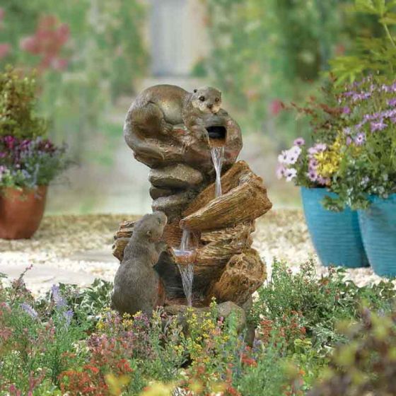 A charming garden scene showcases the La Hacienda Water Fountain - Distinctive with two otter statues from La Hacienda. One otter is perched on top, with water flowing from its mouth, while the other stands below. This distinctive design, surrounded by colorful flowers and vibrant green foliage, is perfect for adding a touch of elegance to your home decor.