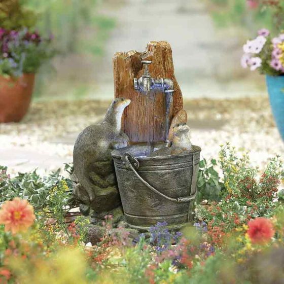 Discover the enchanting allure of the La Hacienda Water Fountain - Distinctive by La Hacienda. This whimsical garden scene features a faux-aged water pump installed on a wooden post, pouring water into a weathered metal bucket. Perched on the bucket are two charming animal figurines: one a beaver and the other possibly a gopher. Surrounding the display, vibrant flowers add color and magic to this delightful garden centerpiece.