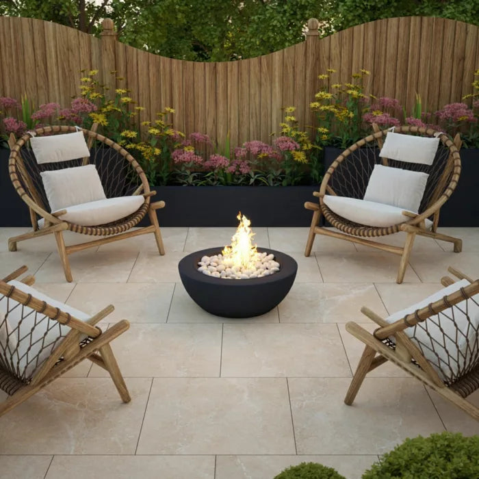 The patio showcases four woven outdoor chairs with white cushions placed around a black fire pit filled with stones. In the background, there's a wooden fence decorated with pink and yellow flowers. The floor is paved with Brisks' Next Bone Porcelain Paving, featuring light stone tiles enhanced by a slip-resistant surface for stylish safety.