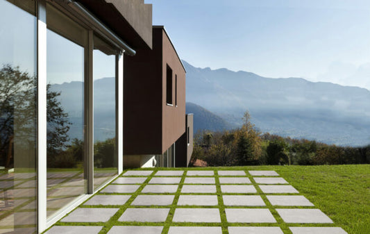 A modern house with expansive glass windows and a tiled patio overlooks a picturesque mountain view. The outdoor area showcases Maestrat Perla Porcelain Paving by Brisks, interspersed with grass to provide a slip-resistant surface. The vibrant landscape includes trees under a clear blue sky.