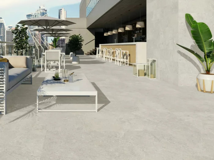 The rooftop terrace showcases modern furniture and elegant design, with white tables, chairs, and a row of bar stools lining the counter. Potted plants and a large umbrella provide shade over Brisks' Next Marfil Porcelain Paving. Enjoy the city skyline as you relax under the clear sky.