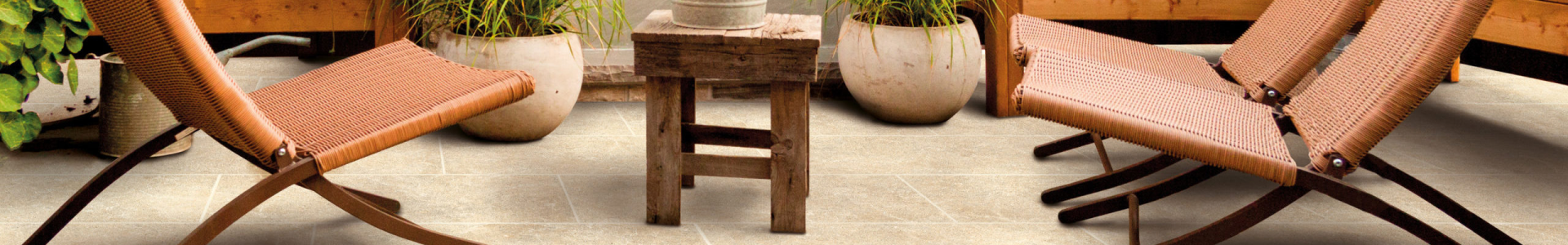 A cozy outdoor seating area features two brown wicker lounge chairs facing each other, a rustic wooden table in the middle, and large ceramic pots with plants in the background. The area is set on a tiled floor.