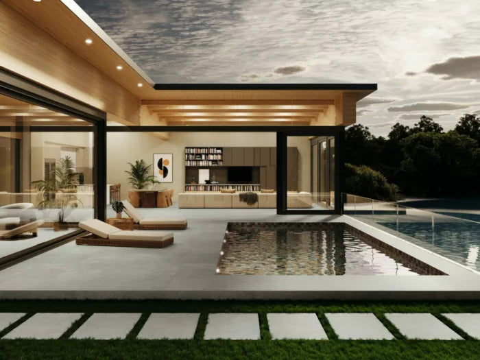 A contemporary home with expansive glass windows, epitomizing urban elegance, offers a view of a reflecting pool adorned with minimalist outdoor furniture. Inside, the cozy living space is enhanced by bookshelves and potted plants. The tranquil atmosphere is beautifully complemented by the overcast sky backdrop against the Brisks Next Grigio Porcelain Paving.