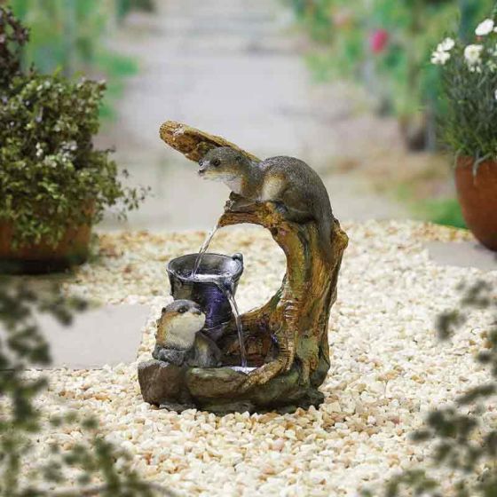 A garden scene with a stone path and potted plants. In the center, the La Hacienda Water Fountain - Distinctive showcases two otter figurines. One otter is perched on a tree trunk pouring water into a jug, while the other otter looks up, catching the water at the base. It's truly one of La Hacienda's distinctive designs in home decor.