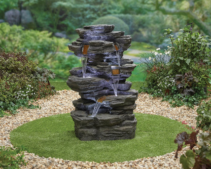 A La Hacienda Hinoki Springs (Inc. LED) Water Feature by La Hacienda stands in the center of a lush garden. Water cascades down its multiple tiers into small pools, subtly illuminated by LED lights and surrounded by a circular area of green grass edged with white pebbles. The background is filled with plants and trees.