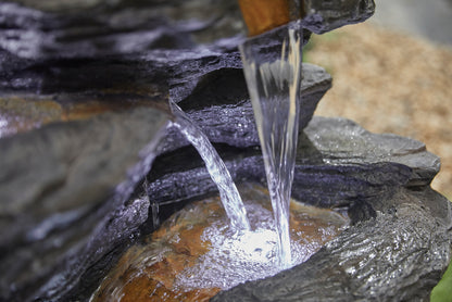 A close-up of the La Hacienda Hinoki Springs (Inc. LED) Water Feature. Water cascades from a wooden spout, flowing over layered rocks and illuminating the base. The tranquil scene, complemented by blurred natural elements in the background, creates a peaceful ambiance reflective of La Hacienda's craftsmanship.