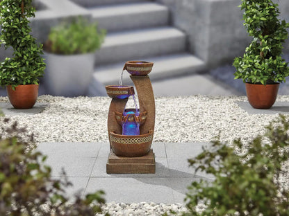 A sophisticated Azure Columns inc LEDS water feature from La Hacienda, crafted from poly-resin and enhanced by LED lighting, adorns the stone patio. Its cascading water shimmers with blue illumination, set amidst planters filled with lush green shrubs and a gravel area that guides you to enchanting stone steps.