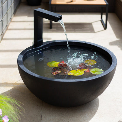 A contemporary La Hacienda Patio Pond features a sleek water element in a stylish black basin, complete with floating lily pads. The water elegantly cascades from a straight spout, generating gentle ripples. This fountain beautifully enhances any outdoor setting and is displayed on a beige-tiled surface with a wooden table in the background.