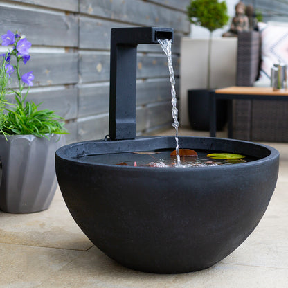 A contemporary La Hacienda Patio Pond in black, featuring a tall spout cascading water into its round basin, crafts a peaceful water feature. Graceful lily pads float atop the surface, while a gray pot nearby holds radiant purple flowers. The tranquil outdoor area is perfected with a wooden fence and stylish patio furniture.