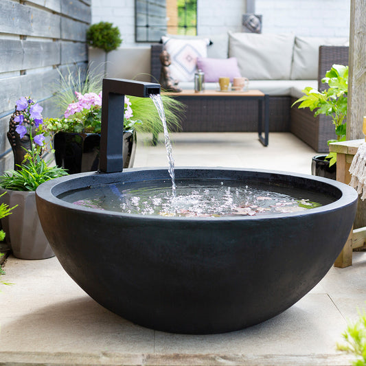 A contemporary outdoor area features La Hacienda's Patio Pond, a substantial black water fountain bowl that generates a tranquil patio pond with its gentle flow. Surrounded by potted plants and a wicker sofa on tiled flooring, the ambiance is peaceful and welcoming.