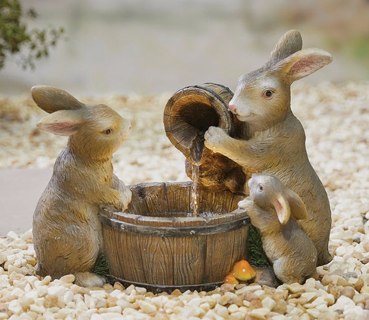 Three beige and brown rabbit figurines gather around a wooden bucket, embodying La Hacienda Water Fountain - Distinctive's unique design. One rabbit pours water from a small barrel into the larger bucket while the other two watch, creating a charming home decor piece. The scene is set on a bed of white pebbles and showcases La Hacienda's distinct craftsmanship.