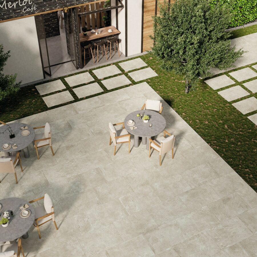 Outdoor patio area with Brisks’ Priorat Bone Porcelain Paving - Individual Tiles, featuring round gray tables and cushioned chairs. The space has a green lawn with stepping stones and a tree. A building with a shaded, wooden entrance labeled "Café" is visible in the background.