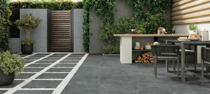 This contemporary outdoor patio highlights slip-resistant Priorat Graphite Porcelain Paving by Brisks and square stepping stones surrounded by gravel. A wooden gate and a vibrant green wall add a charming backdrop, while the dining area includes a table with chairs and a countertop decorated with logs and kitchen items.