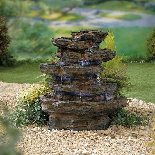 A La Hacienda Red Rock Springs (Inc. LED) Water Feature, featuring water cascading down multiple levels of a sturdy resin structure, is surrounded by small pebbles and greenery. This fountain, enhanced by subtle LED lighting, is set against a backdrop of a garden with blurred foliage and a glimpse of a green field in the distance.