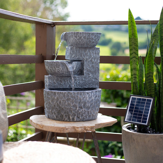 A charming solar water feature, the Pouring Bowls by La Hacienda, graces the patio, with water elegantly cascading through its tiered stone bowls. It rests on a wooden table beside a potted plant with long green leaves, and to its right, a small solar panel powers this delightful piece.