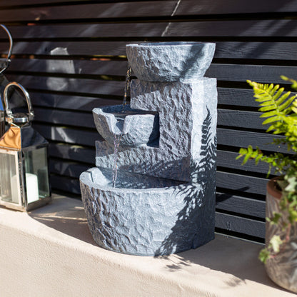 The La Hacienda Pouring Bowls fountain elegantly adorns a ledge against a dark slatted wall. With water flowing smoothly from the top bowl to the bottom, it is powered by a solar water feature system. A nearby decorative lantern adds charm to the scene, accompanied by a lush potted fern.