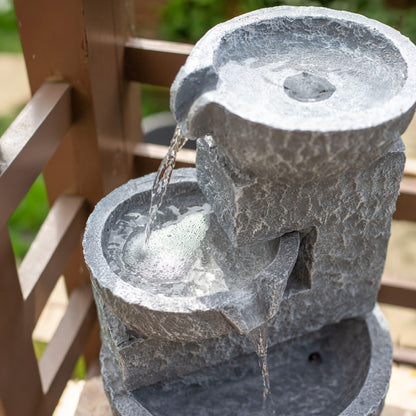 A close-up view of the Pouring Bowls by La Hacienda, a two-tiered stone effect water fountain that is self-contained and elegant. Water gracefully cascades from the top bowl into the lower one, creating gentle ripples. The fountain is set against a wooden lattice background, offering a harmonious blend of nature and artistry.