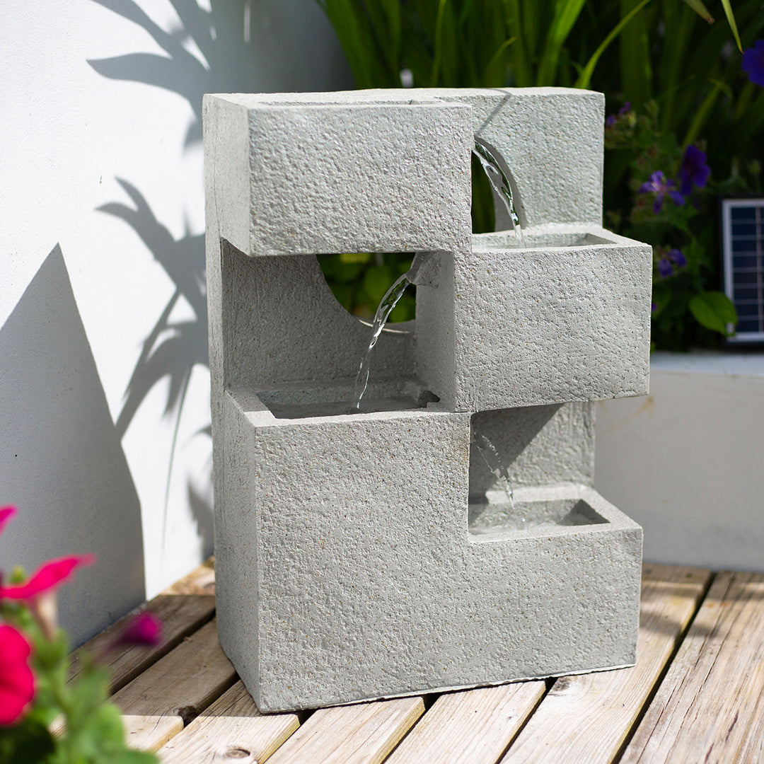 Cube Falls by La Hacienda showcases a concrete-effect design with cascading streams in a minimalist style, gracefully positioned on a wooden deck. Enveloped by greenery and flowers, this solar-powered water feature incorporates a small solar panel in the background for an eco-friendly touch.