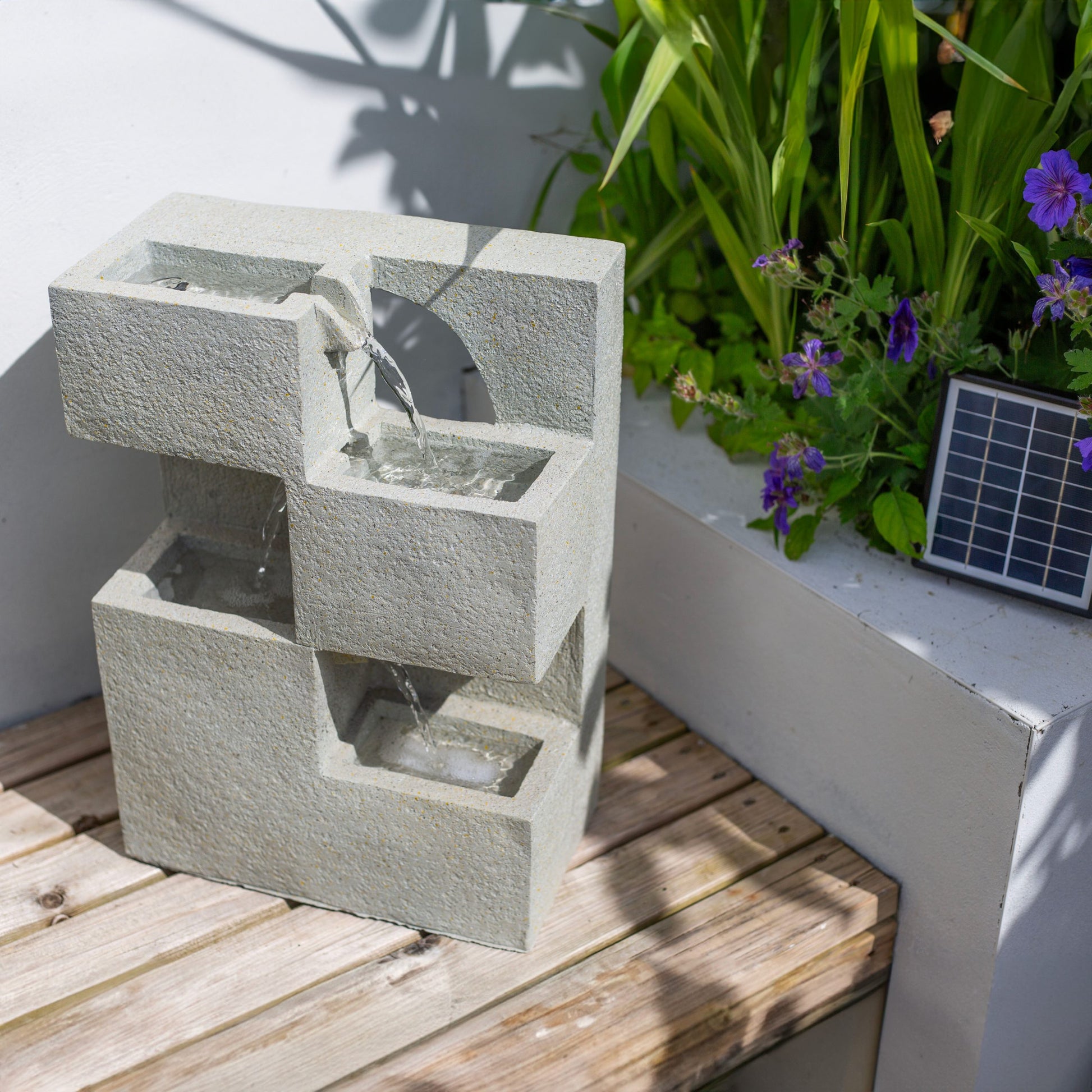 A La Hacienda Cube Falls water feature, designed with a geometric shape and cascading water, rests on a wooden bench. It is surrounded by lush green plants and vibrant purple flowers, presenting a concrete-effect design. A small solar panel nearby powers this eco-friendly centerpiece.