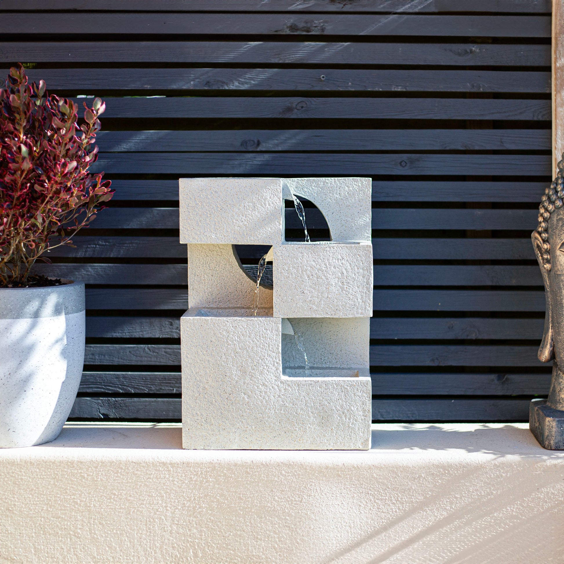 The Cube Falls by La Hacienda is a contemporary tabletop solar-powered water feature characterized by its geometric square shapes, allowing water to cascade through its central cutouts. Its concrete-effect design is ideally set on a ledge, accented with a decorative plant on the left and partially framed by a statue on the right.