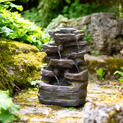 The Rock Falls by La Hacienda, a compact solar-powered fountain, features cascading water and is designed to be surrounded by lush green moss and plants in a garden setting, offering a tranquil and natural ambiance.