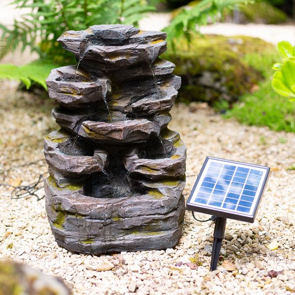 The Rock Falls, a compact, solar-powered water feature by La Hacienda, is designed to cascade water down stacked rocks into a base pool. It sits on gravel while being powered by a solar panel mounted on a nearby stake and is surrounded by lush greenery and mossy stones.