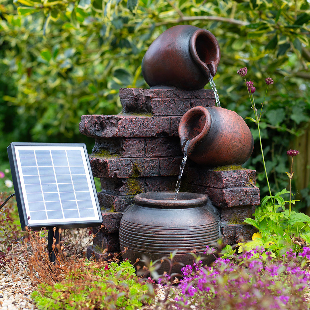 The La Hacienda Pot Wall Pours is a solar-powered water feature showcasing two large clay pots positioned on a brick structure, with water elegantly flowing from one to another below. It stands amidst small purple flowers and lush foliage, enhancing the garden's tranquility and outdoor ambiance.