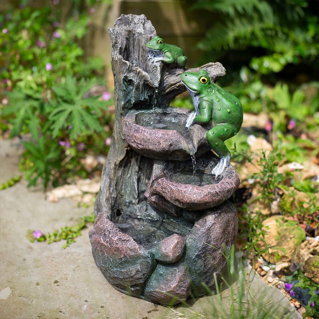 A garden scene showcasing two lifelike green frog sculptures from La Hacienda's Frog Pools collection, perched on a solar-powered water fountain. Water gently cascades down the tiered levels, surrounded by lush greenery and delicate purple flowers. Sunlight illuminates this enchanting garden accessory, enhancing the natural ambiance.