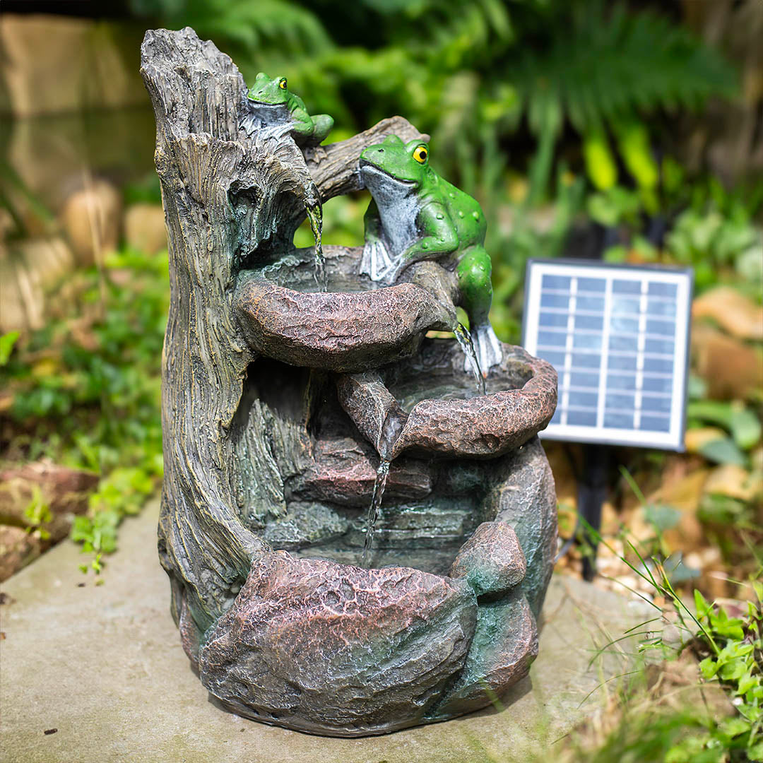 The delightful Frog Pools garden accessory by La Hacienda showcases a tree stump design with two resin frogs sitting on the edge of a solar water fountain. Water flows gracefully down several tiers, and an unobtrusive solar panel harmoniously integrates into the surrounding lush greenery.