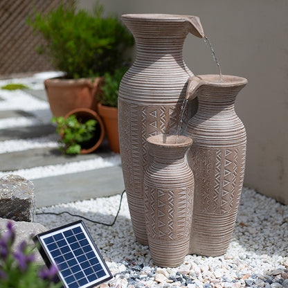 The Aztec Vase by La Hacienda features three decorative, textured clay jugs artistically stacked in descending order, allowing water to flow from one to the next and creating a serene garden oasis. A small solar panel powers this captivating feature, which is set amidst pebbles and potted plants for a harmonious natural backdrop.