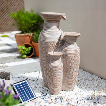 La Hacienda's Aztec Vase, a solar-powered water feature, transforms a garden into a serene retreat. This stunning piece features three beige ceramic vases with intricate patterns that cascade delicately, reminiscent of traditional Aztec designs. Set against white gravel and surrounded by vibrant greenery, the subtle presence of a discreet solar panel maintains its peaceful ambiance without interruption.