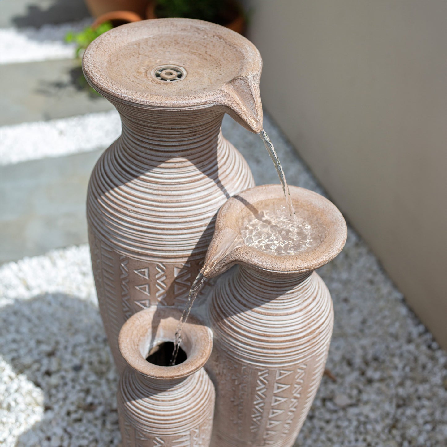 La Hacienda's Aztec Vase features a three-tier design with a tribal pattern, forming a solar-powered water feature that elegantly cascades water from the top vase to the lower ones. Set on a bed of white pebbles and surrounded by potted plants, this fountain creates a serene garden oasis against a light-colored wall.