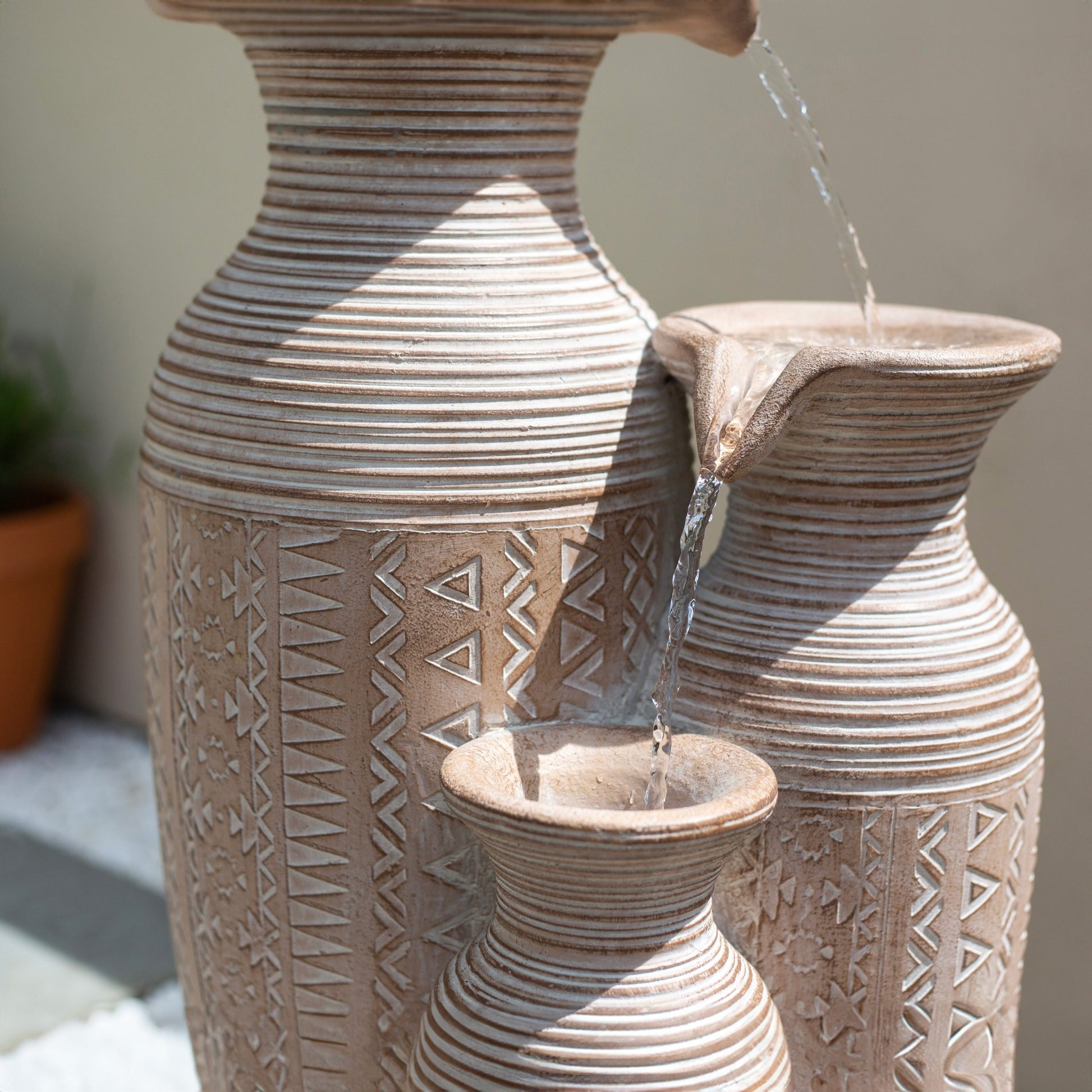 A decorative water feature is formed by three intricately patterned clay pitchers, with the design reminiscent of La Hacienda's Aztec Vase. Powered by solar energy, the sunlight accentuates their texture, while a small potted plant adds to this garden oasis in the background.