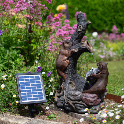 A garden scene featuring the Woodland Otters by La Hacienda: a solar-powered water feature designed to resemble a tree trunk. Two intricately crafted squirrels and otters rest on the trunk while water cascades from above. A discreet solar panel is surrounded by vibrant flowers and lush greenery.