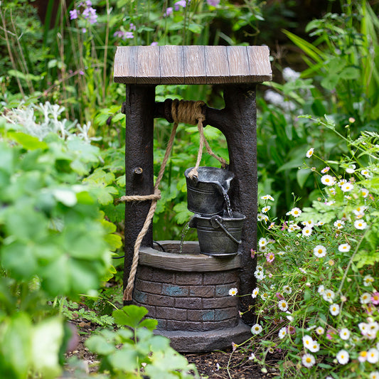 The Wishing Well incl LEDs by La Hacienda features a vintage-style design with a wooden roof and rope, complemented by a metal bucket filled with water. This charming garden decoration is enhanced by lush greenery and wildflowers surrounding it.