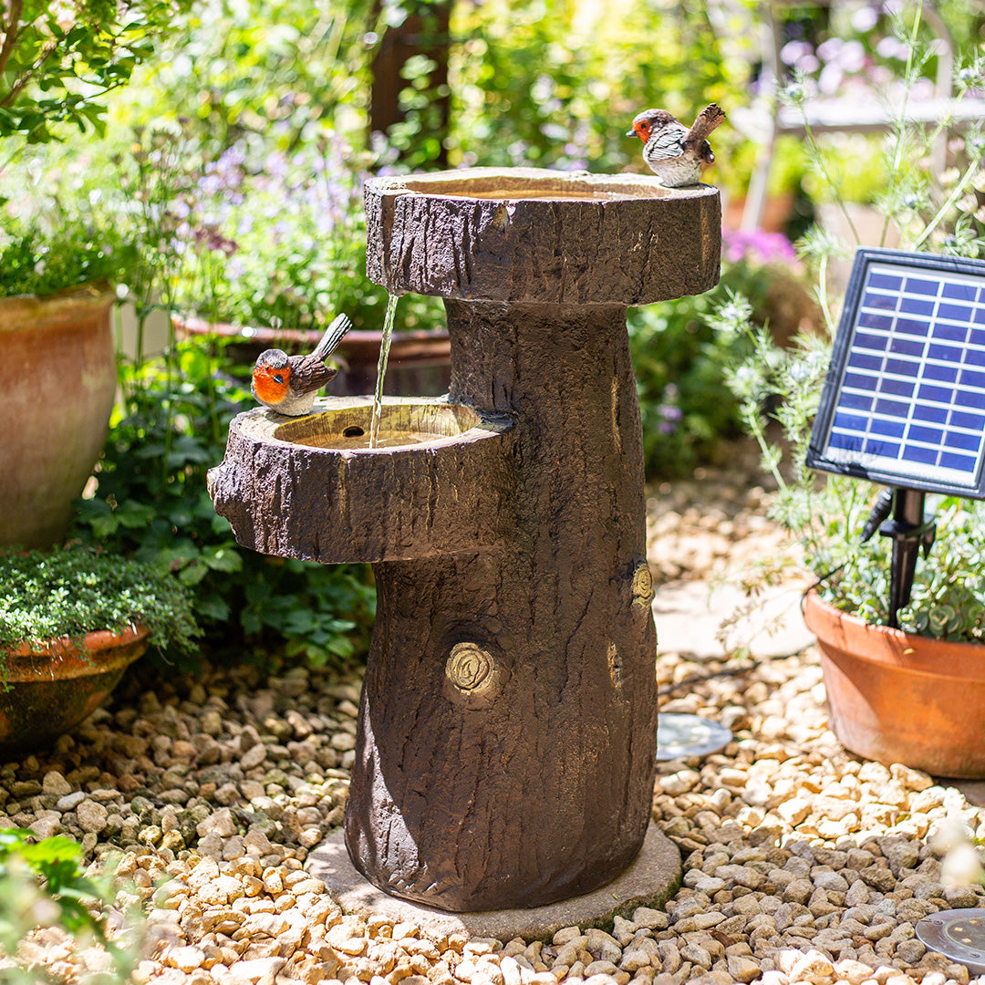 Birds perched on the Robin Springs incl LEDs, a solar-powered bird bath fountain designed to resemble a rustic tree trunk, nestled among potted plants and pebbles. A small solar panel sparkles on the right, highlighting this La Hacienda garden water feature in its sunlit setting.